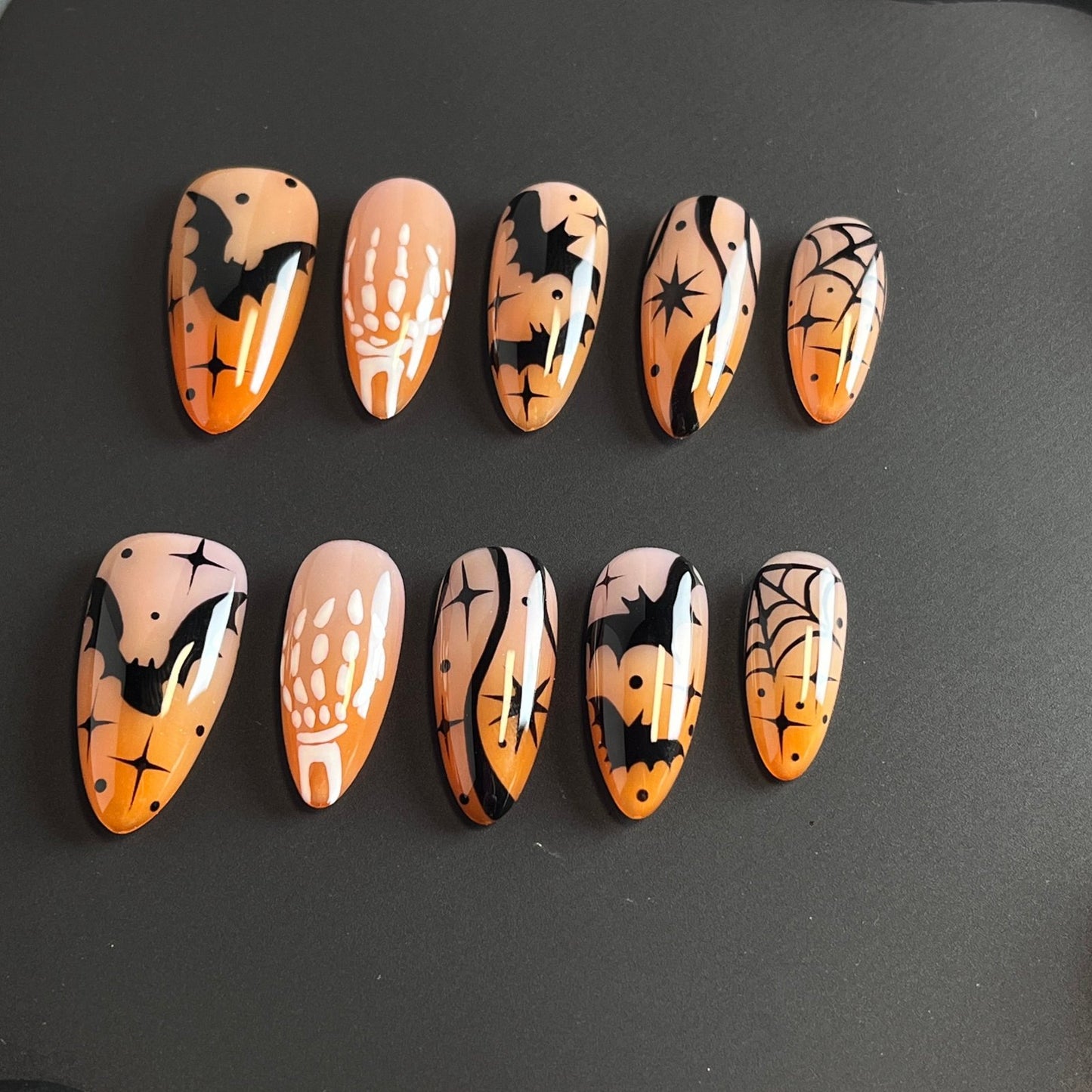 Haunted Nightfall press on nails, featuring hauntingly beautiful designs of bats, skeleton hands, spider webs, and cosmic stars over a soft nude-orange gradient